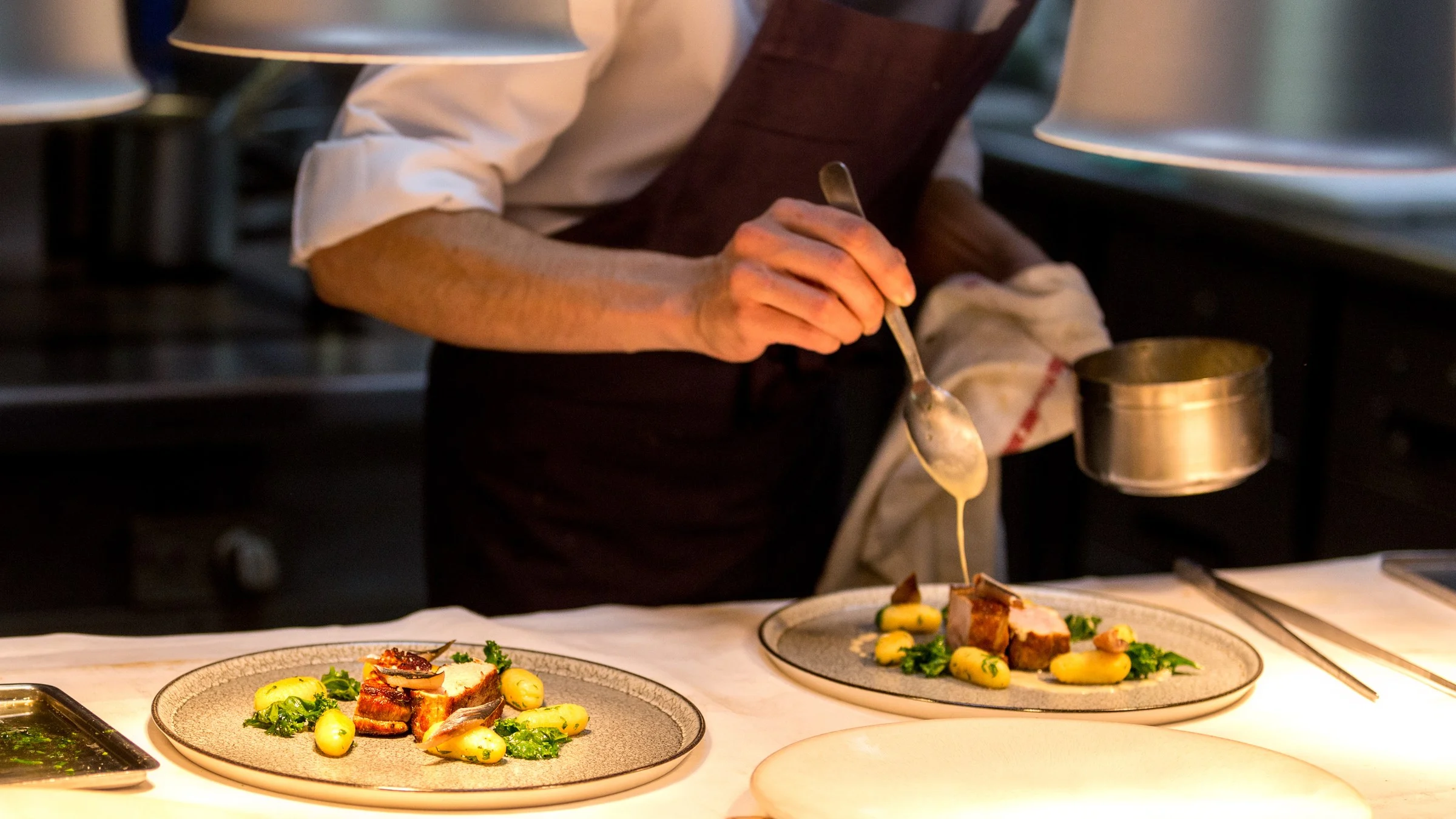 A chef plating two dishes on the pass in a commercial kitchen.