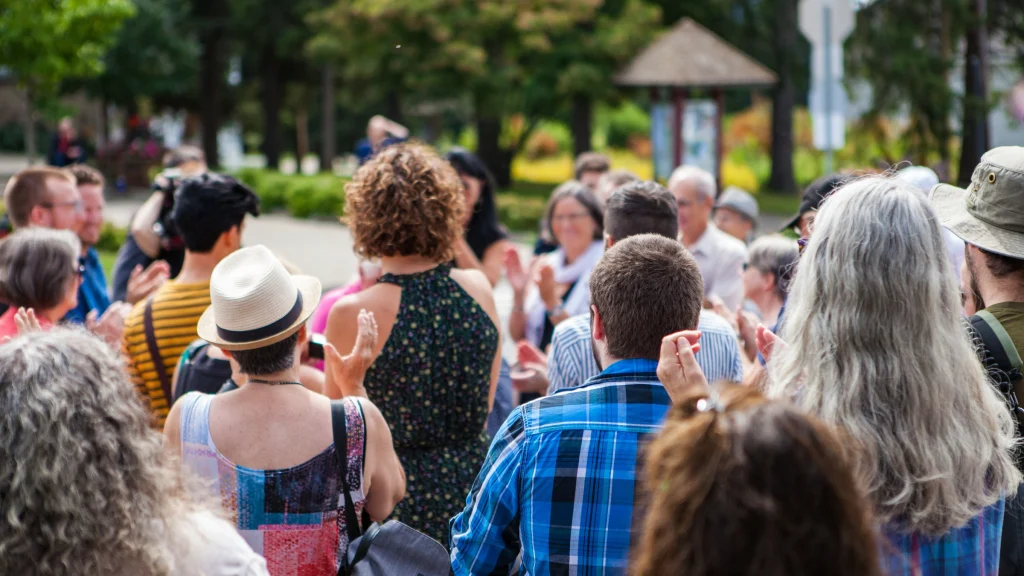 A community crowd gathered outdoors, applauding, seen from behind.